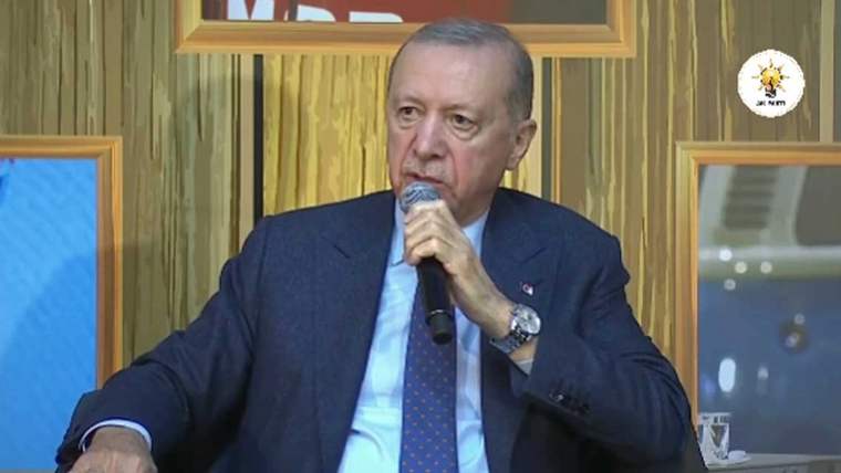 Bir kişi, mikrofonla konuşma yapıyor, arka planda ahşap görünümlü bir duvar var.