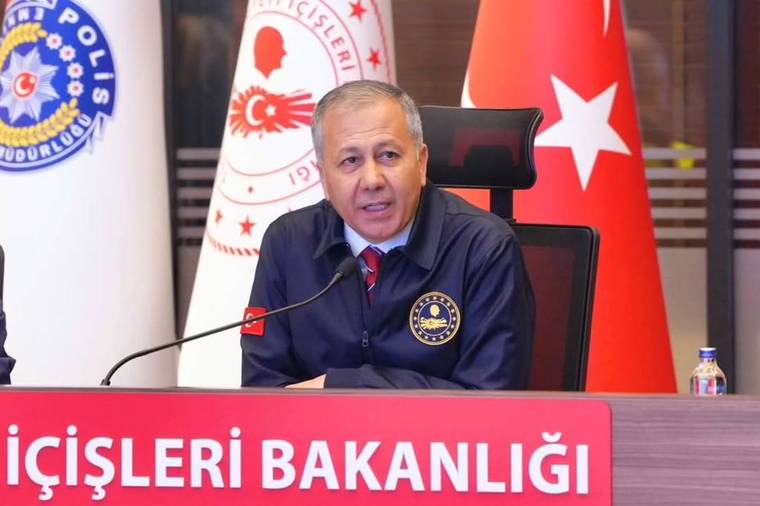 Kürsüde oturan bir kişi mikrofona konuşuyor; arkasında Türk bayrakları ve İçişleri Bakanlığı amblemleri bulunuyor.