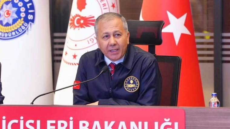 Kürsüde oturan bir kişi mikrofona konuşuyor; arkasında Türk bayrakları ve İçişleri Bakanlığı amblemleri bulunuyor.