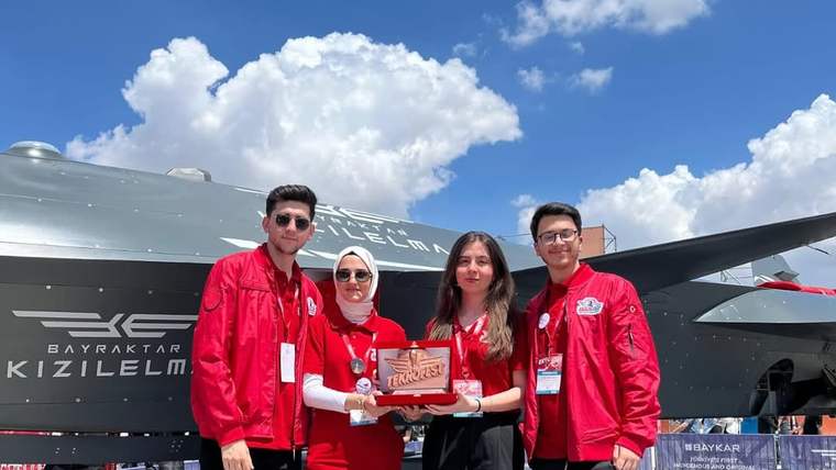 Teknofest alanında dört kişi, uçak ve mühimmat maketi önünde ellerinde plaketle duruyor.