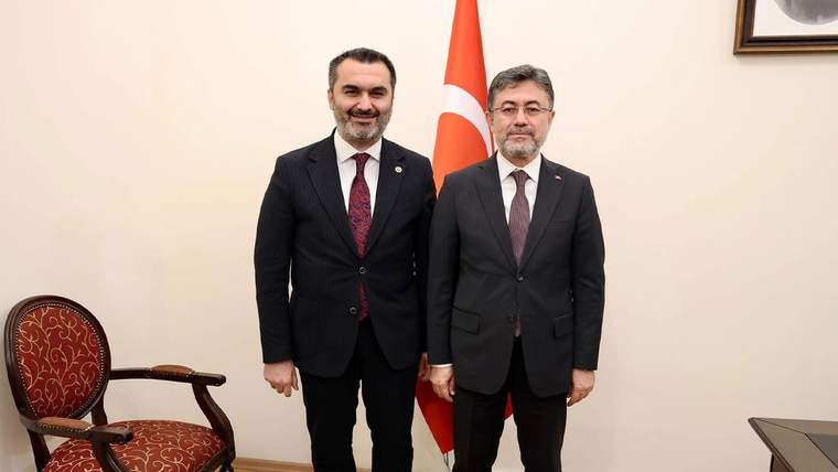 Türk bayrağının önünde takım elbiseli iki kişi, bir odada yan yana ayakta duruyor.