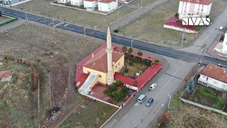 Kırmızı çatılı, tek minareli bir cami; çevresinde yollar, evler, araçlar ve boş araziler görülüyor.