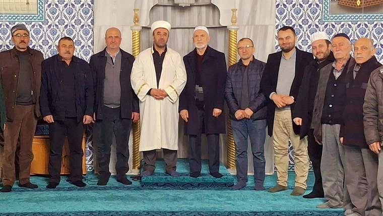 Cami içinde, mihrap önünde farklı yaşlarda 11 erkek yan yana duruyor; zeminde mavi halı görülüyor.