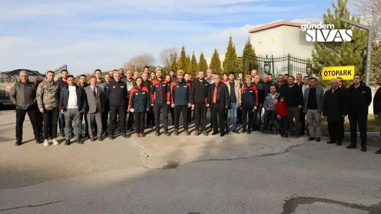 Bir grup insan, otopark önünde topluca poz veriyor. Arka planda ağaçlar ve bir bina görünmekte.