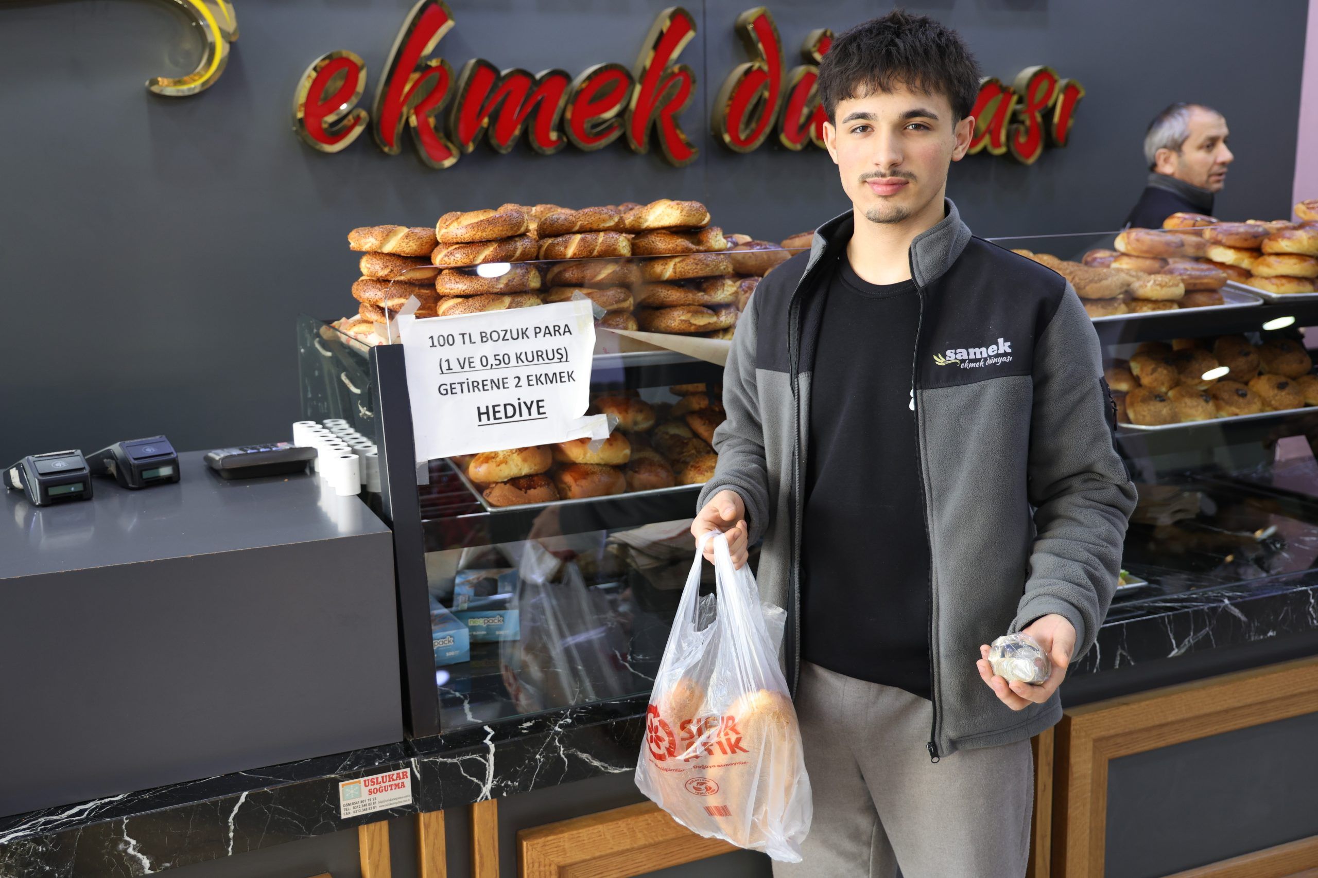 Fırın tezgâhında ekmekler sergilenirken genç bir kişi elinde poşet ve bozuk paralarla duruyor.