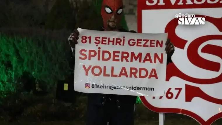 Örümcek maskeli bir kişi, üzerinde “81 şehri gezen Spiderman yollarda” yazan tabela tutuyor.