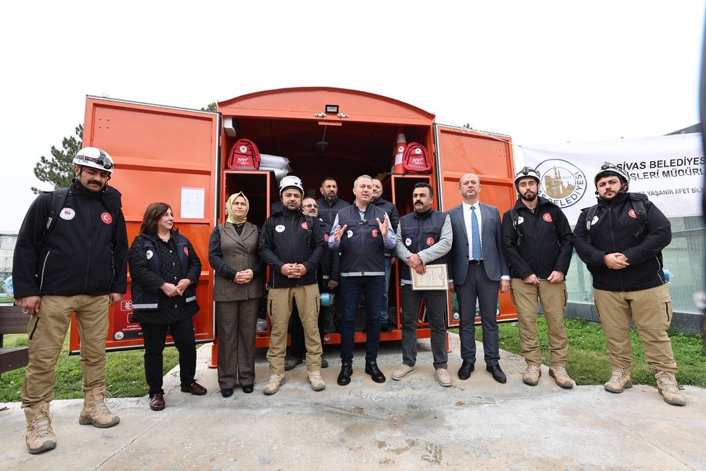 Açık turuncu konteynerin önünde, aralarında üniformalı kişilerin de bulunduğu bir grup poz veriyor.