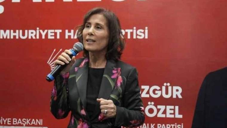 Mikrofon tutan bir kadın, kırmızı fonda parti yazıları önünde konuşurken el hareketi yapıyor.