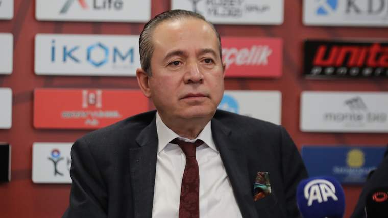 Mikrofonlar önünde oturan takım elbiseli bir kişi, sponsor logolarının bulunduğu fonda konuşuyor.