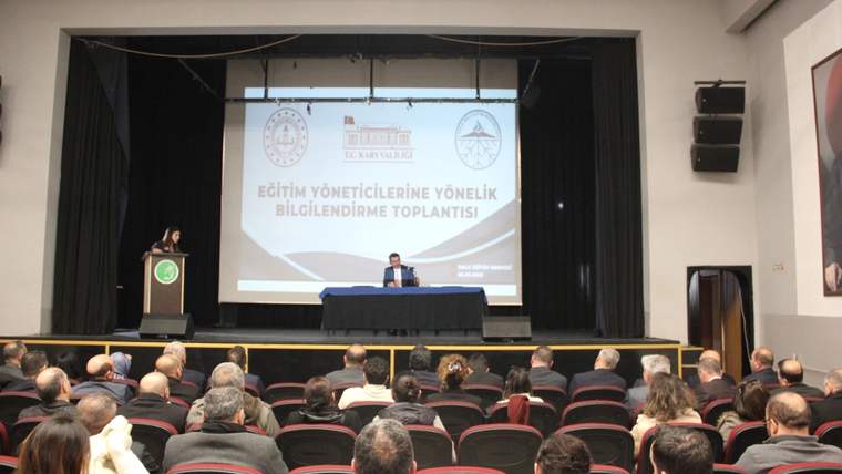 Salonda oturan izleyiciler, sahnedeki kürsü ve masanın arkasında toplantı başlığı bulunan perdeyi izliyor.