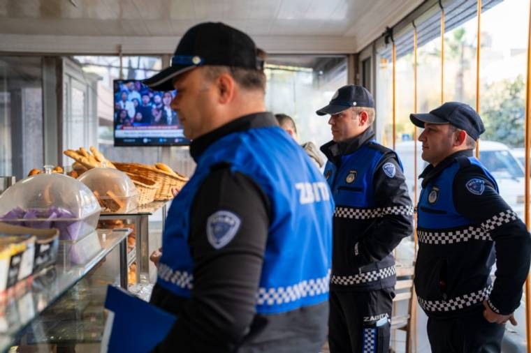 Bir iş yerinde üç zabıta görevlisi, cam tezgâhtaki ekmek ve hamur işlerinin yanında duruyor.