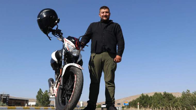 Motosikletinin yanında duran bir adam, açık mavi gökyüzü altında gülümsüyor.