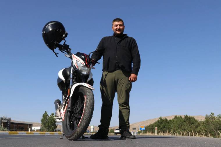 Motosikletinin yanında duran bir adam, açık mavi gökyüzü altında gülümsüyor.