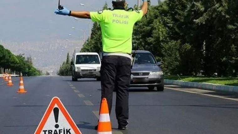 Trafik kontrolü yapan bir polis, yolda durdurma işareti yapıyor; önünde trafik kontrol tabelası ve koni var.
