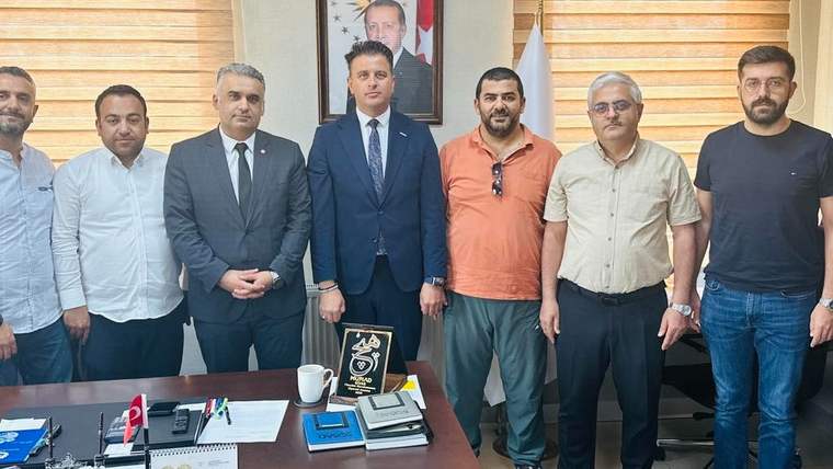 Bir grup erkek, bir ofiste yan yana duruyor; masada belgeler ve bir ödül var.