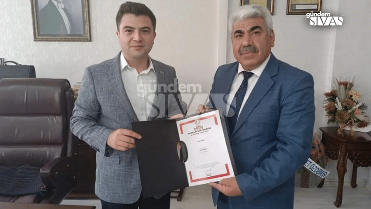 Bir kişi, elinde belge tutarken diğer kişiyle birlikte poz veriyor.
