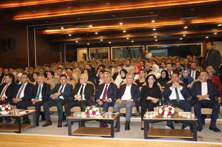 Toplantı salonunda takım elbiseli ve günlük giyimli çok sayıda kişi sıralar halinde oturuyor.