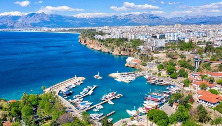 Kıyı boyunca uzanan turkuaz deniz, yat limanı, yoğun yerleşim ve arka planda dağlar görülüyor.