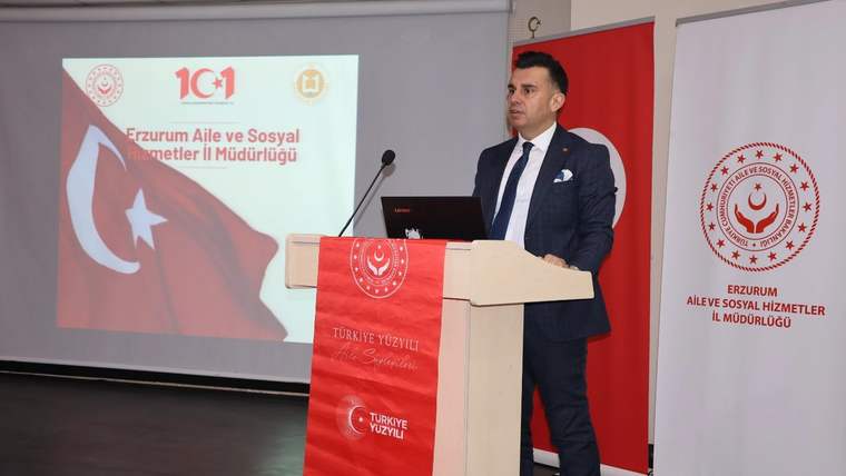 Kürsüde konuşan bir kişi, arkasındaki ekranda Türk bayrağı ve kurum adı bulunan salonda duruyor.