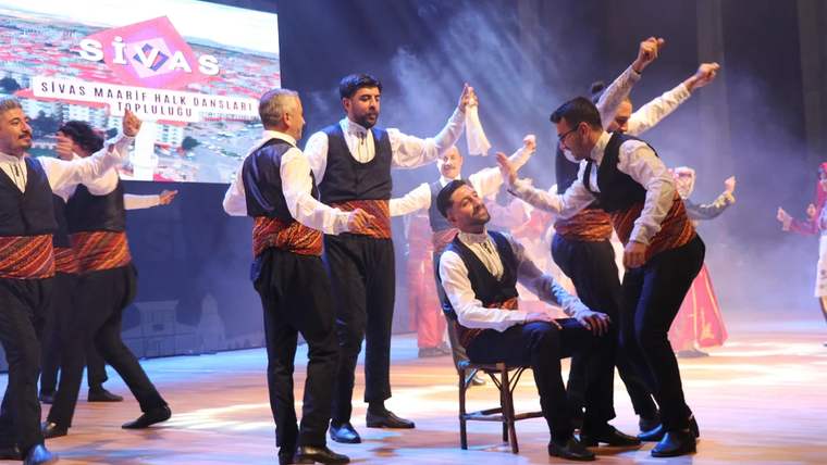 Sahne üzerinde halk oyunları sergileyen dansçılar, geleneksel kıyafetler giymekte.