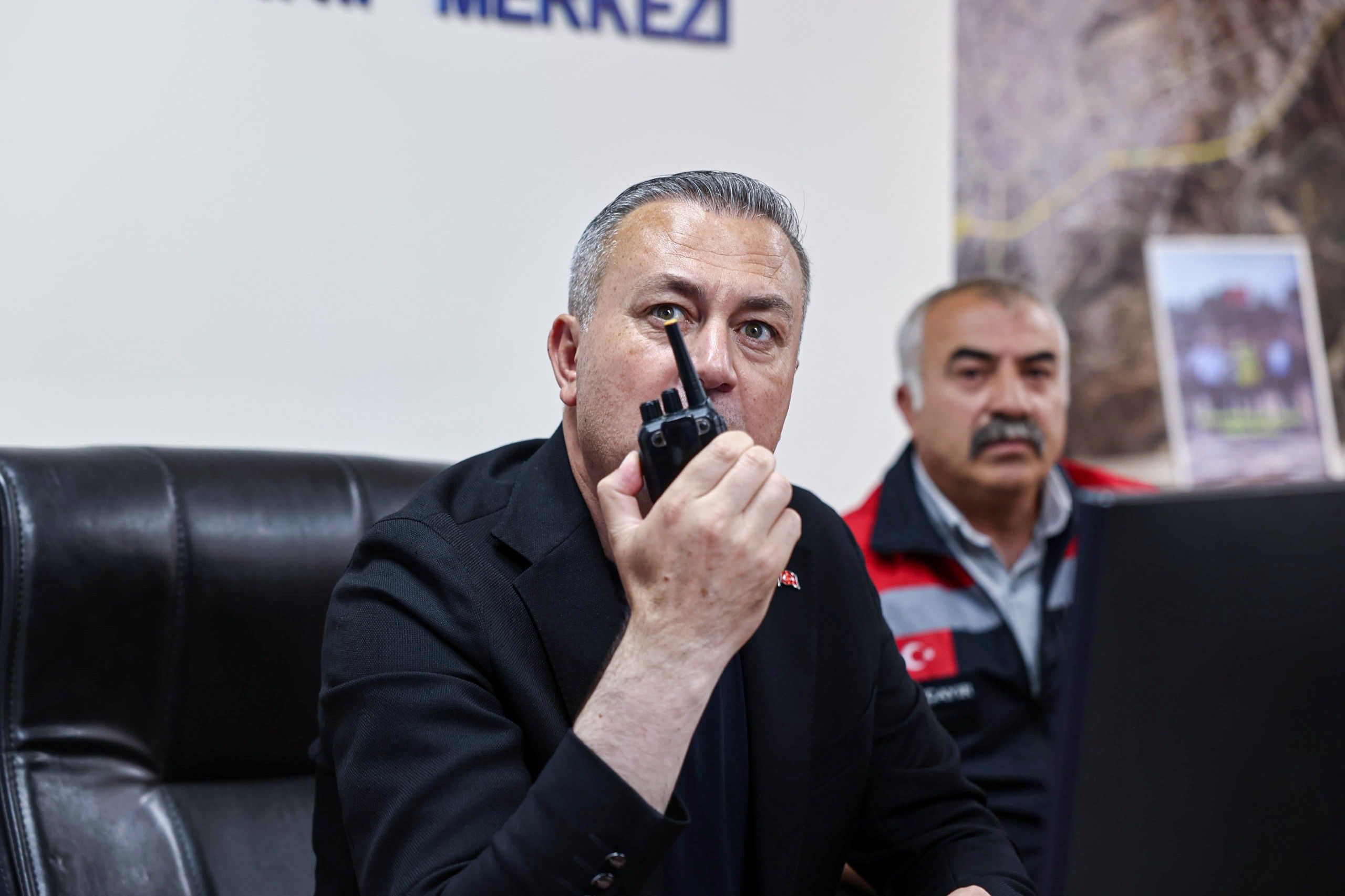 Siyah montlu bir kişi elindeki telsizle konuşurken, arkasında kırmızı yelekli başka bir kişi oturuyor.