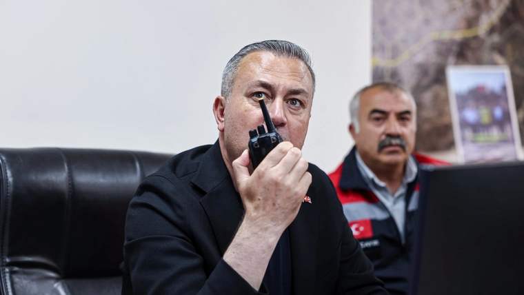 Siyah montlu bir kişi elindeki telsizle konuşurken, arkasında kırmızı yelekli başka bir kişi oturuyor.