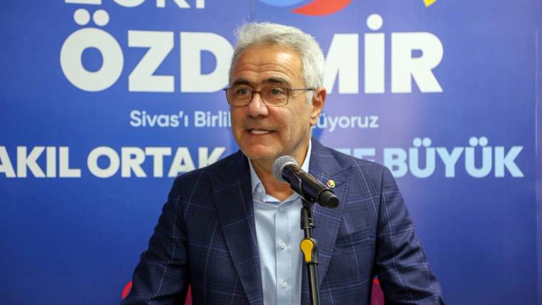 Mikrofon karşısında konuşan gözlüklü bir kişi, üzerinde “ÖZDEMİR” yazan mavi pankart önünde duruyor.
