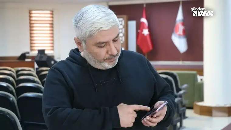 Beyaz saçlı bir kişi, toplantı salonunda elindeki telefona bakarken parmağıyla ekranı işaret ediyor.