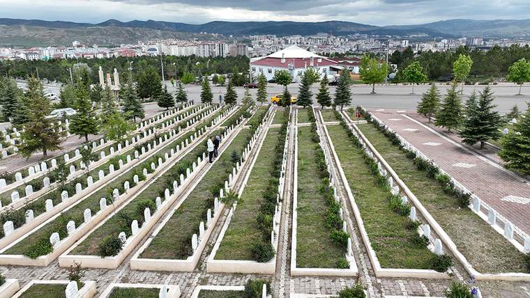Bir mezarlıkta düzenli sıralanmış mezar taşları, arka planda şehir manzarası ve dağlar görünmekte.