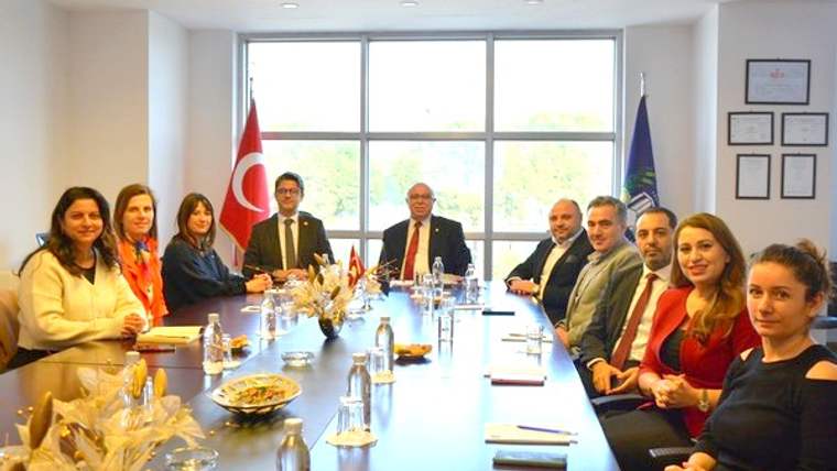 Bir toplantı odasında, masanın etrafında oturan dokuz kişi ve arka planda Türk bayrağı ile kurum bayrağı görünmekte.