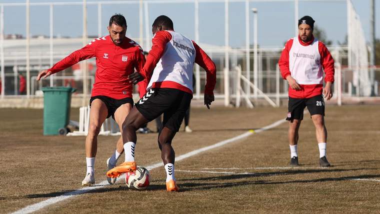 İki futbolcu, antrenman sahasında pas çalışması yapıyor; arka planda başka oyuncular ve antrenman ekipmanları var.