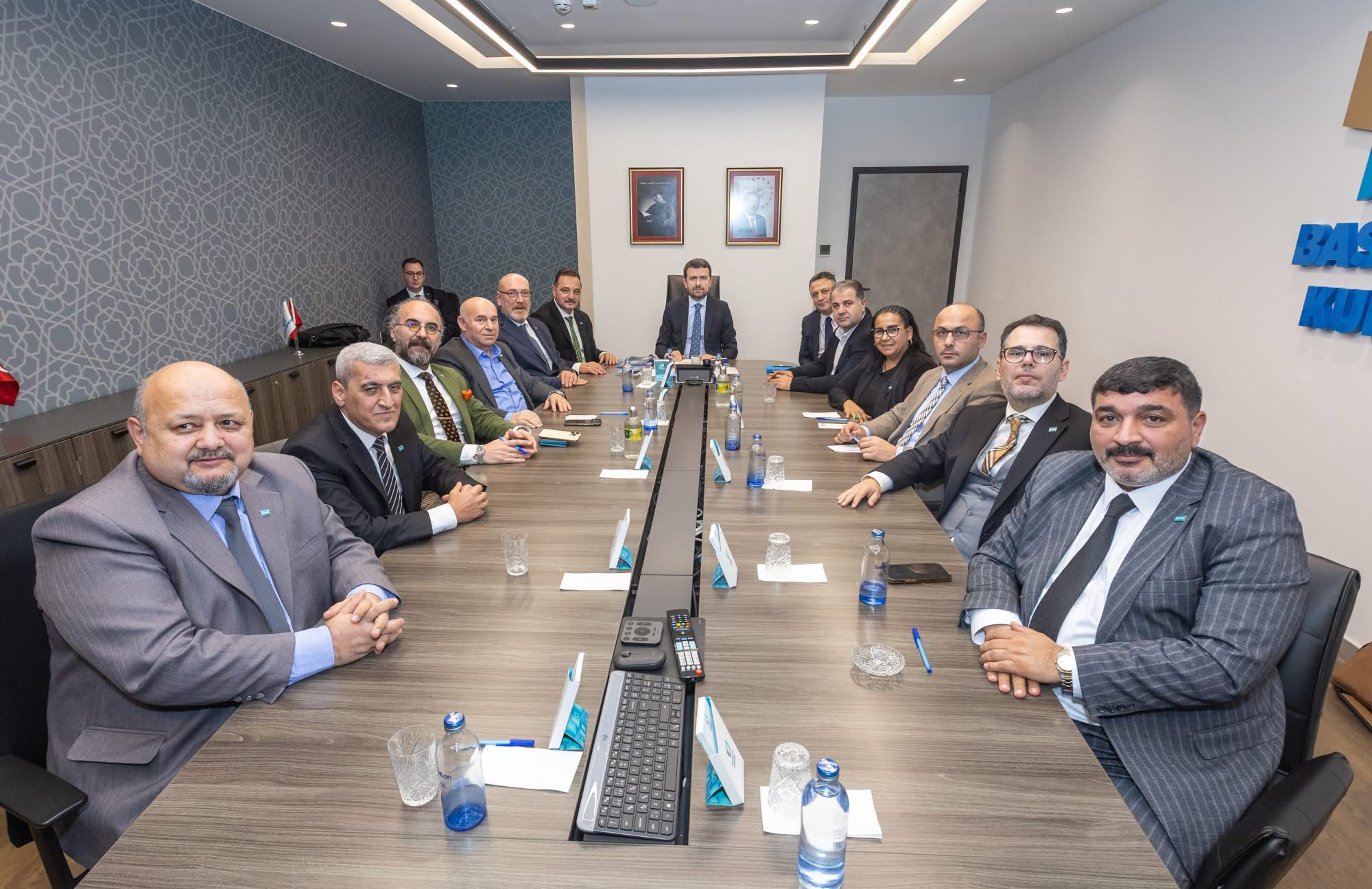 Toplantı odasında uzun masa çevresinde takım elbiseli kişiler oturuyor; masada mikrofonlar ve su şişeleri bulunuyor.