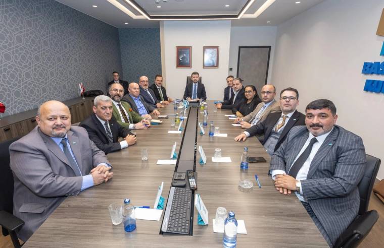 Toplantı odasında uzun masa çevresinde takım elbiseli kişiler oturuyor; masada mikrofonlar ve su şişeleri bulunuyor.