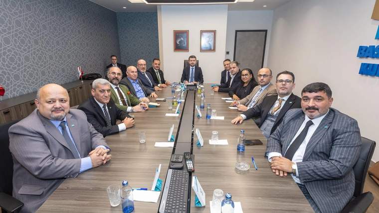 Toplantı odasında uzun masa çevresinde takım elbiseli kişiler oturuyor; masada mikrofonlar ve su şişeleri bulunuyor.