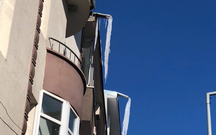 Bir apartmanın balkon ve saçaklarından aşağıya uzanan uzun buz sarkıtları, açık mavi gökyüzü önünde görülüyor.