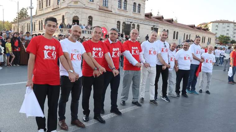 Kırmızı ve beyaz tişört giyen kişiler, tarihi taş binanın önünde el ele sıra oluşturuyor.