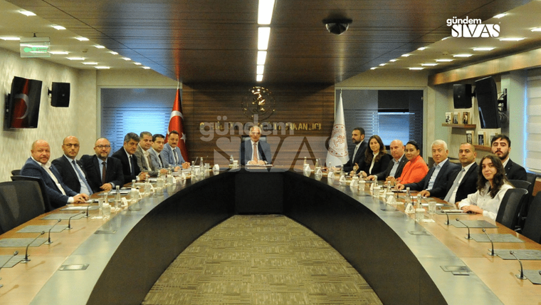 Bir toplantı odasında, masanın etrafında oturan çok sayıda kişi ve masanın üzerinde içecekler var.