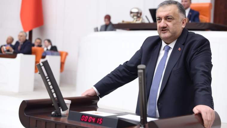 Kürsüde konuşan bir kişi, önünde zaman sayacı ve mikrofon var. Arkada koltuklarda başka kişiler oturuyor.
