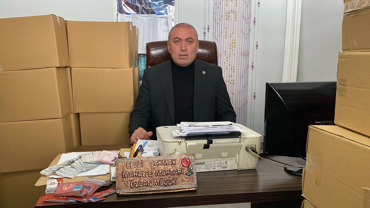 Masa başında oturan bir kişi, çevresindeki koliler ve evraklarla birlikte kameraya bakıyor.