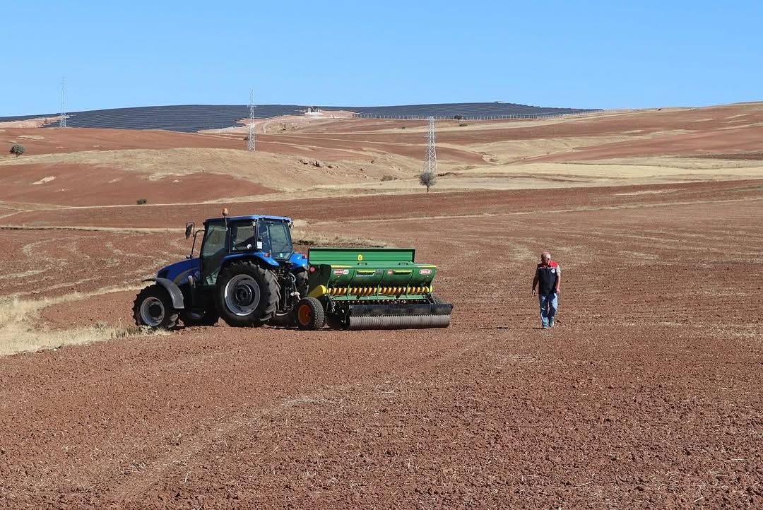 Kırmızı toprakta bir traktör ve ekim makinesi, yanında yürüyen bir kişi görünüyor.