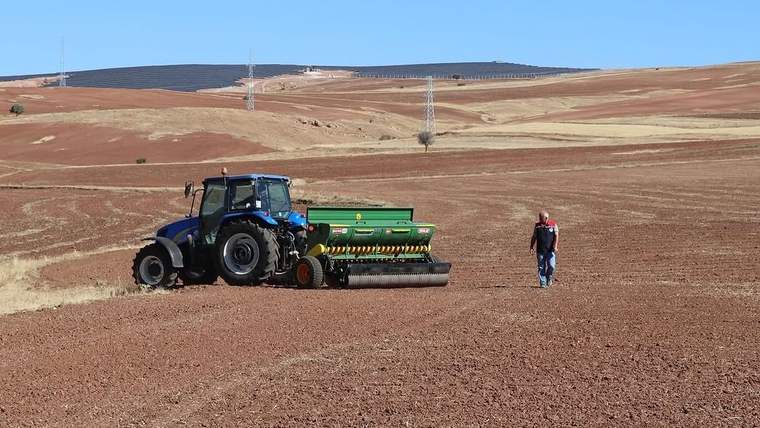 Kırmızı toprakta bir traktör ve ekim makinesi, yanında yürüyen bir kişi görünüyor.