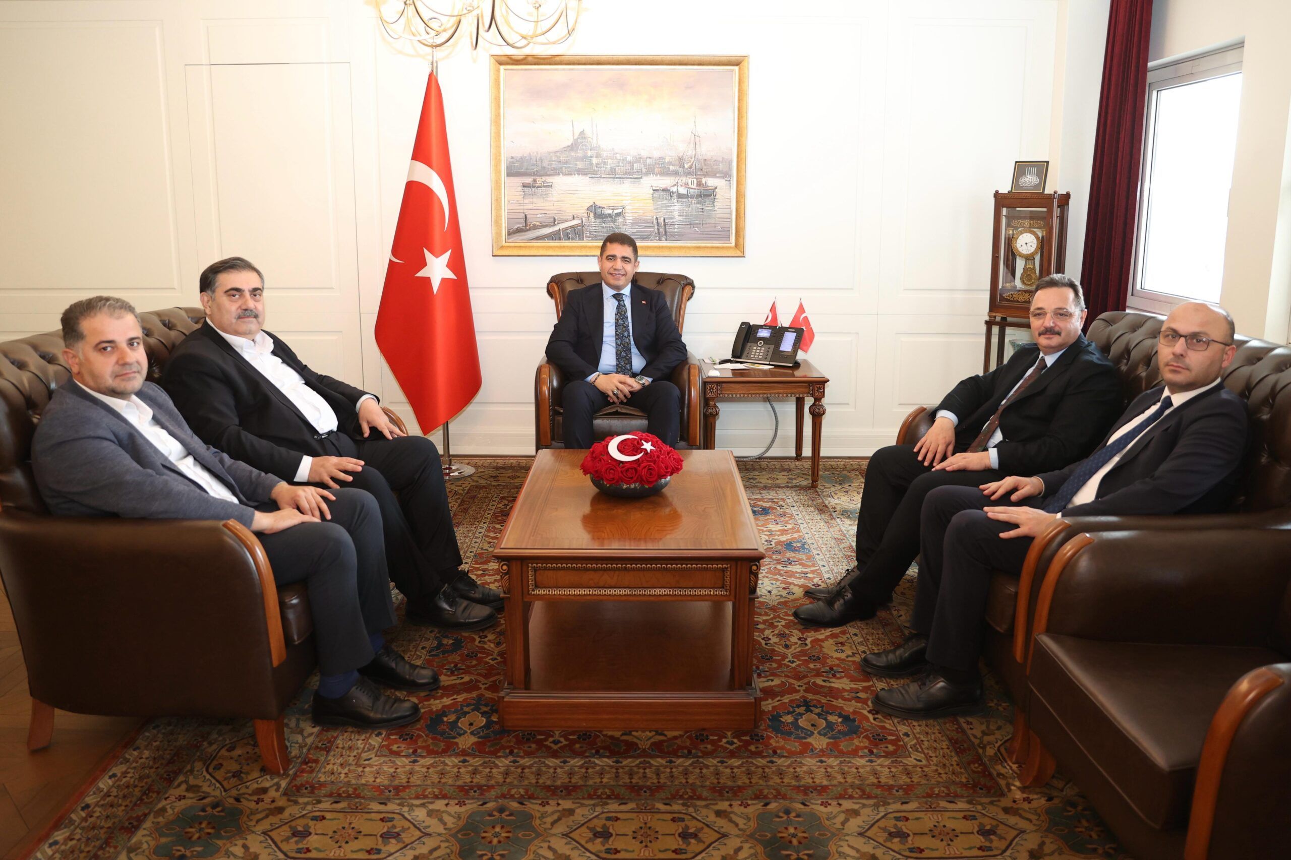 Türk bayrağının bulunduğu odada, sehpa çevresinde oturan altı erkek görülüyor.