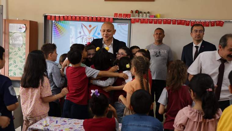 Sınıfta çocuklar, öğretmenler ve yetişkinler bir ekranın önünde toplanmış; duvarlarda Türk bayrakları asılı.