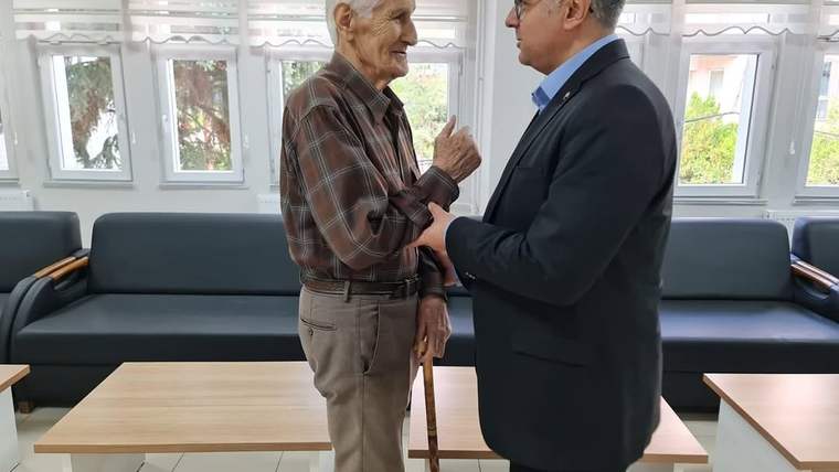 Baston taşıyan yaşlı bir erkek, yanında duran takım elbiseli erkekle aydınlık bir oturma alanında konuşuyor.