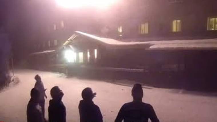 Gece karla kaplı alanda aydınlatılmış bir bina ve önünde duran dört kişinin silüeti görülüyor.