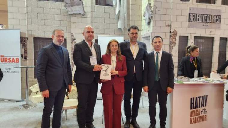 Bir fuar standı önünde beş kişi poz verirken, ortadaki kadın elinde bir broşür tutuyor; tezgahta Hatay yazıyor.