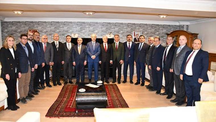 Bir salonda, çoğu takım elbiseli çok sayıda kişi duvar önünde yan yana duruyor; ortada sehpa ve halı bulunuyor.
