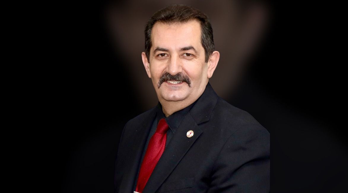 Koyu arka planda, takım elbise giymiş bir adam gülümseyerek poz veriyor.