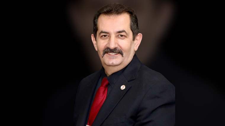 Koyu arka planda, takım elbise giymiş bir adam gülümseyerek poz veriyor.