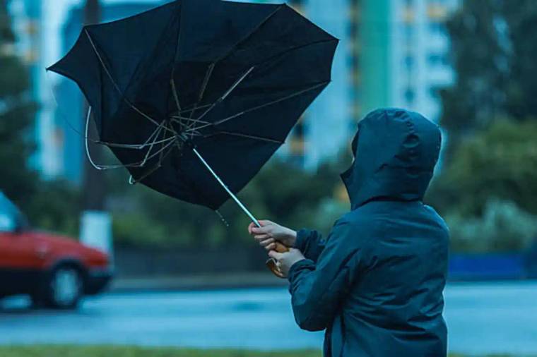 Yağmurluklu bir kişi, rüzgârda ters dönmüş siyah şemsiyeyi tutarken ıslak yol görünüyor.
